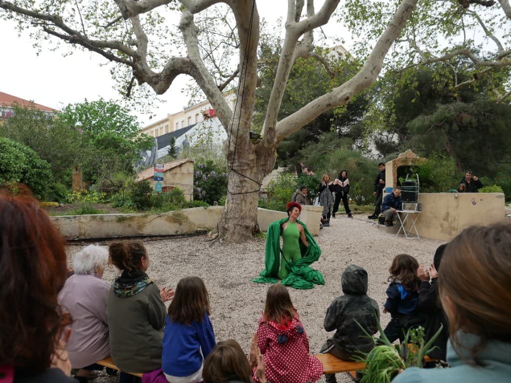 Performance artistique en costume vert devant un public assis, sous un grand platane dans la cour du jardin.