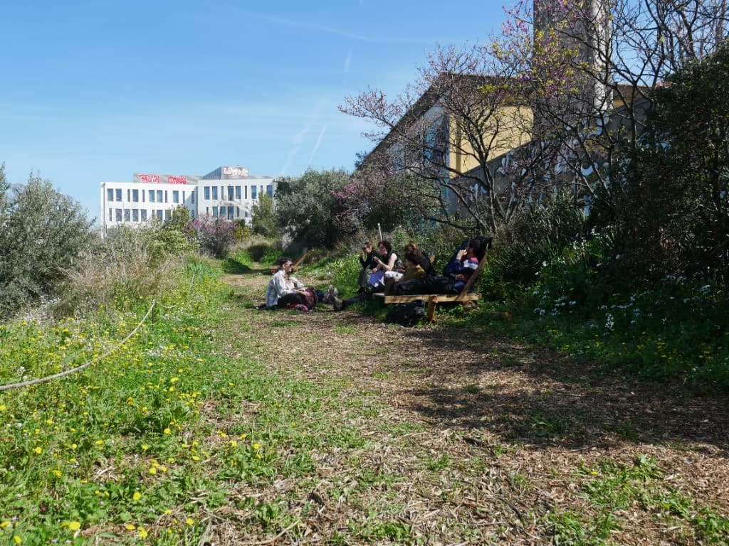 Groupe de personnes assises sur des bancs en bois le long d'un chemin couvert de copeaux, dans la verdure du jardin.